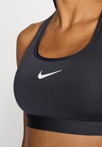 Μαύρο αθλητικό σουτιέν από ελαστικό ύφασμα, με λαιμόκοψη σε σχήμα scoop και λευκό λογότυπο Nike στο κέντρο του στήθους. Λείο υλικό χωρίς σχέδια.