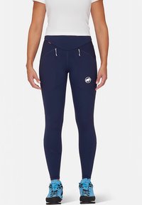 Granatowe legginsy z elastycznego materiału, wyposażone w wysoki pas, kieszenie na zamek oraz logo na lewym udzie. Połączone z niebieskim obuwiem.