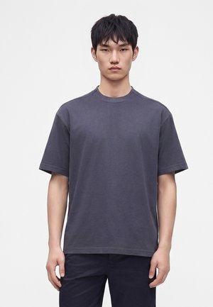 Jeune homme aux cheveux courts et noirs portant un t-shirt gris foncé uni à manches courtes et un pantalon sombre, debout devant un fond blanc.