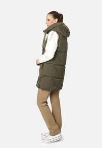 Chaleco acolchado verde con capucha, presenta costuras cuadradas y bolsillos con cremallera en los laterales, llevado sobre una camisa blanca de manga larga y pantalones beige. Zapatillas blancas.