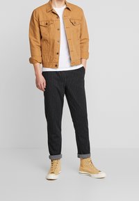 Brun denimjacka över en vit T-shirt, i kombination med svarta randiga byxor och beigefärgade hög-top sneakers. Avslappnad design och relaxed fit.