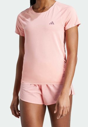 Žena v světle růžovém sportovním tričku s krátkými rukávy a odpovídajících šortkách s malým logem na hrudi, stojící před jednobarevným pozadím.