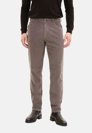 Uomo che indossa pantaloni di velluto a coste color taupe a taglio dritto, con camicia nera a maniche lunghe e stivali in pelle marrone scuro, in piedi davanti a uno sfondo bianco.