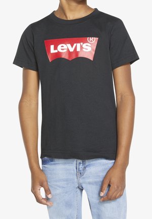 Jongen draagt een zwart Levi's t-shirt met een rood-wit logo, gecombineerd met lichtblauwe spijkerbroeken, tegen een witte achtergrond.
