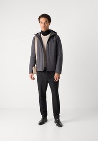 Giacca imbottita grigia con cappuccio, dettagli neri, indossata su un maglione beige. Pantaloni neri e scarpe in pelle completano l'outfit. Borsa a tracolla casual.