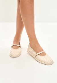 Scarpe basse intrecciate beige con punta rotonda, dotate di una sottile striscia e fibbia, una texture morbida e una suola flessibile.