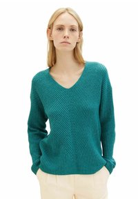 Teal, strukturierter Pullover mit V-Ausschnitt, langen Ärmeln und einem diagonalen Rippenmuster. Über cremefarbenen Hosen getragen.