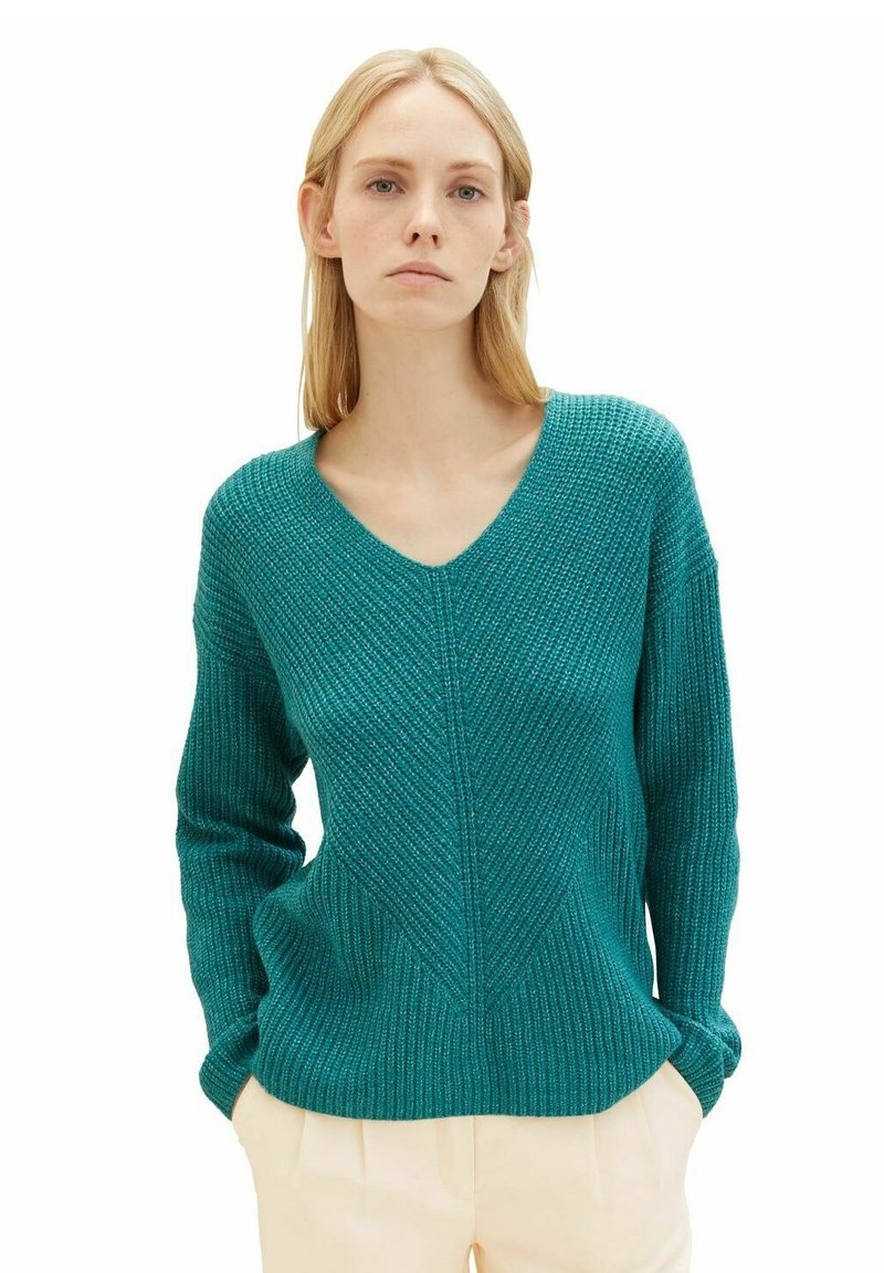 Teal, strukturierter Pullover mit V-Ausschnitt, langen Ärmeln und einem diagonalen Rippenmuster. Über cremefarbenen Hosen getragen.