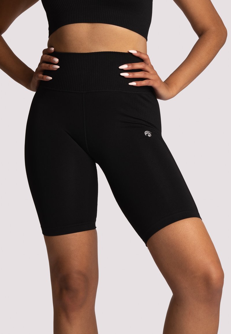 Žena v černých cyklistických šortkách s vysokým pasem a odpovídajícím černým crop topem, s rukama opřenýma o boky na světlém pozadí.