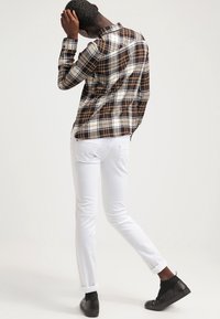 Camisa de franela a cuadros en tonos marrón, blanco y naranja, combinada con jeans blancos ajustados y zapatillas negras. Vista desde atrás.