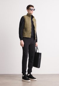 Colete acolchoado verde-oliva por cima de uma blusa de manga longa preta, combinado com calças de jogging pretas e ténis pretos, segurando uma bolsa tote preta.