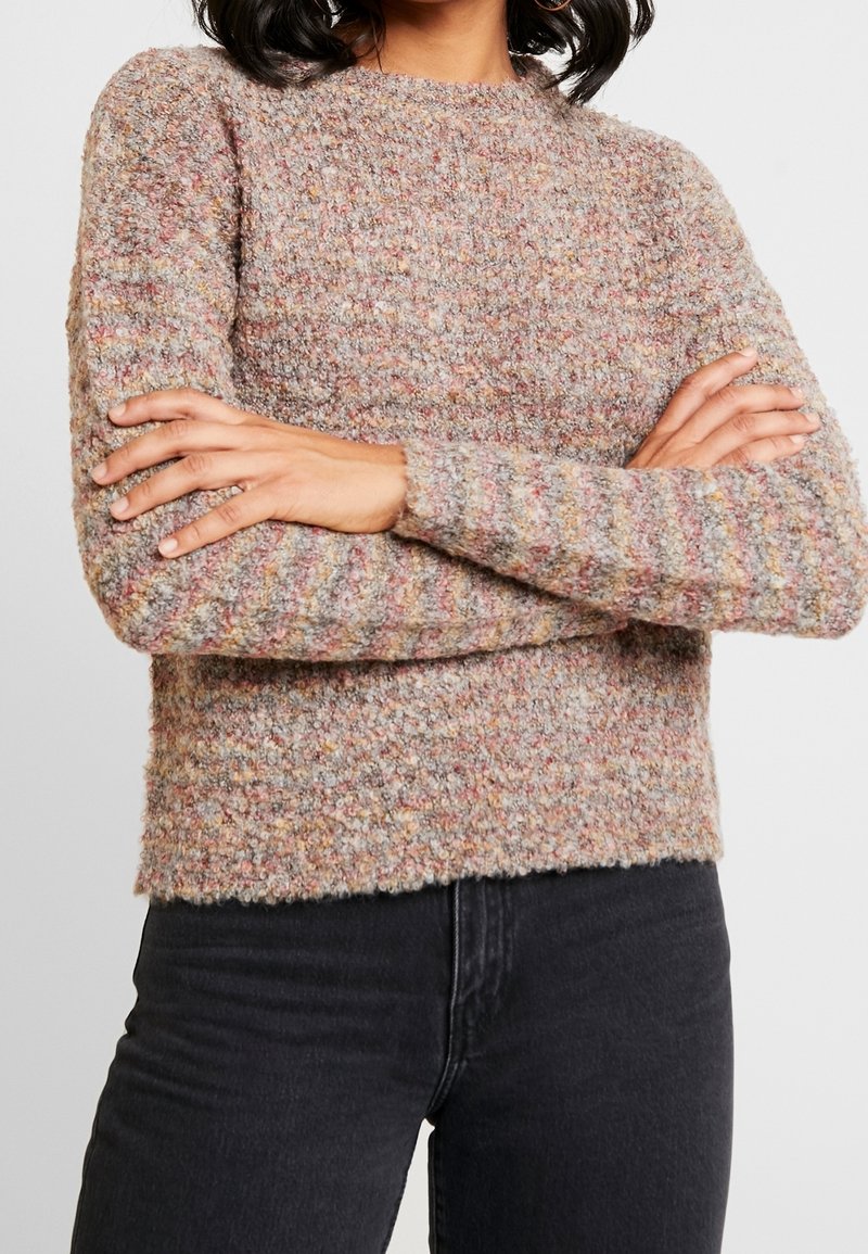 Personne portant un pull en maille multicolore texturé à manches longues, les bras croisés, associé à un jean noir taille haute.