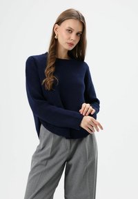 Pull en maille bleu marine avec col rond et épaules tombantes, associé à un pantalon plissé gris clair. Texture douce, coupe décontractée.