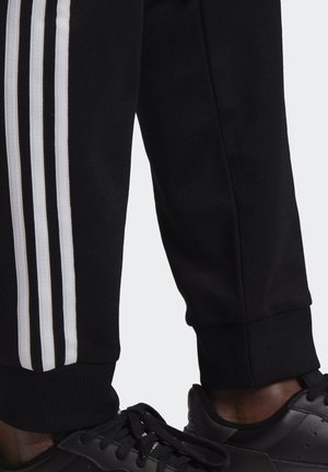 Un pantalon de sport noir présente trois rayures verticales blanches sur les côtés, des poignets côtelés et un tissu doux. Associé à des baskets noires.
