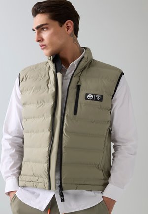 Joven con tatuaje lateral lleva un chaleco acolchado color oliva claro sobre una camisa blanca, mirando hacia su izquierda sobre un fondo liso.