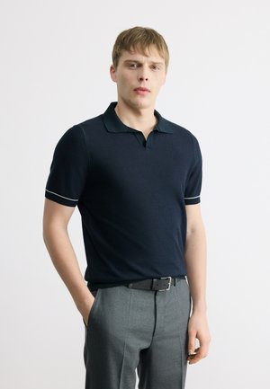 Ung mand iført mørkeblå kortærmet polo og grå bukser står med den ene hånd i lommen mod en ensfarvet baggrund.