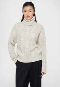 Pull à col roulé en tricot torsadé gris clair, à coupe ample et ourlets côtelés. Le tissu semble doux et texturé. Associé à un pantalon sombre à rayures.