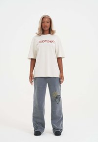 Beige oversized t-shirt med rødt logo, parret med løse grå bukser med broderede designs og dekorativ syning. Sorte fodtøj.