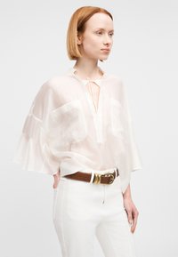 Mujer con cabello rojo corto, vistiendo una blusa blanca translúcida y pantalones blancos con un cinturón ancho marrón con hebilla dorada, con las manos en los bolsillos.