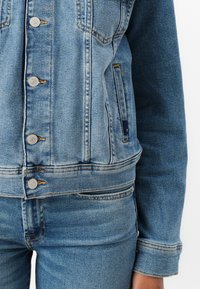 Jachetă din denim de culoare albastru deschis, cu nasturi metalici argintii, două buzunare frontale și o croială ajustată. Prezintă detalii de cusătură și un design clasic al gulerului.
