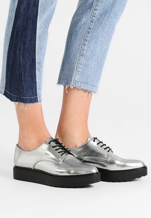 Personne portant des chaussures en cuir métallisé argenté à lacets avec des semelles épaisses noires et un jean bleu bicolore court aux ourlets effilochés.