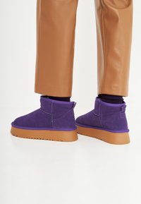 Bottines en suède violet avec une épaisse semelle en caoutchouc brun, ornées de coutures et d'un tirer à l'arrière. Portées avec un pantalon en cuir brun.