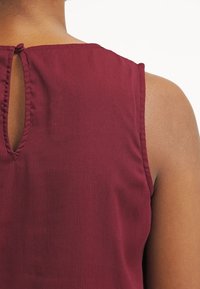Top senza maniche bordeaux in tessuto testurizzato, con una piccola apertura sul retro e dettagli di scollatura arruffata. Materiale morbido e leggero.