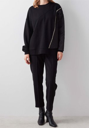 Personne portant un pantalon noir et un haut noir à manches longues avec une couture diagonale et une manche droite matelassée soulignée par une fermeture éclair métallique.