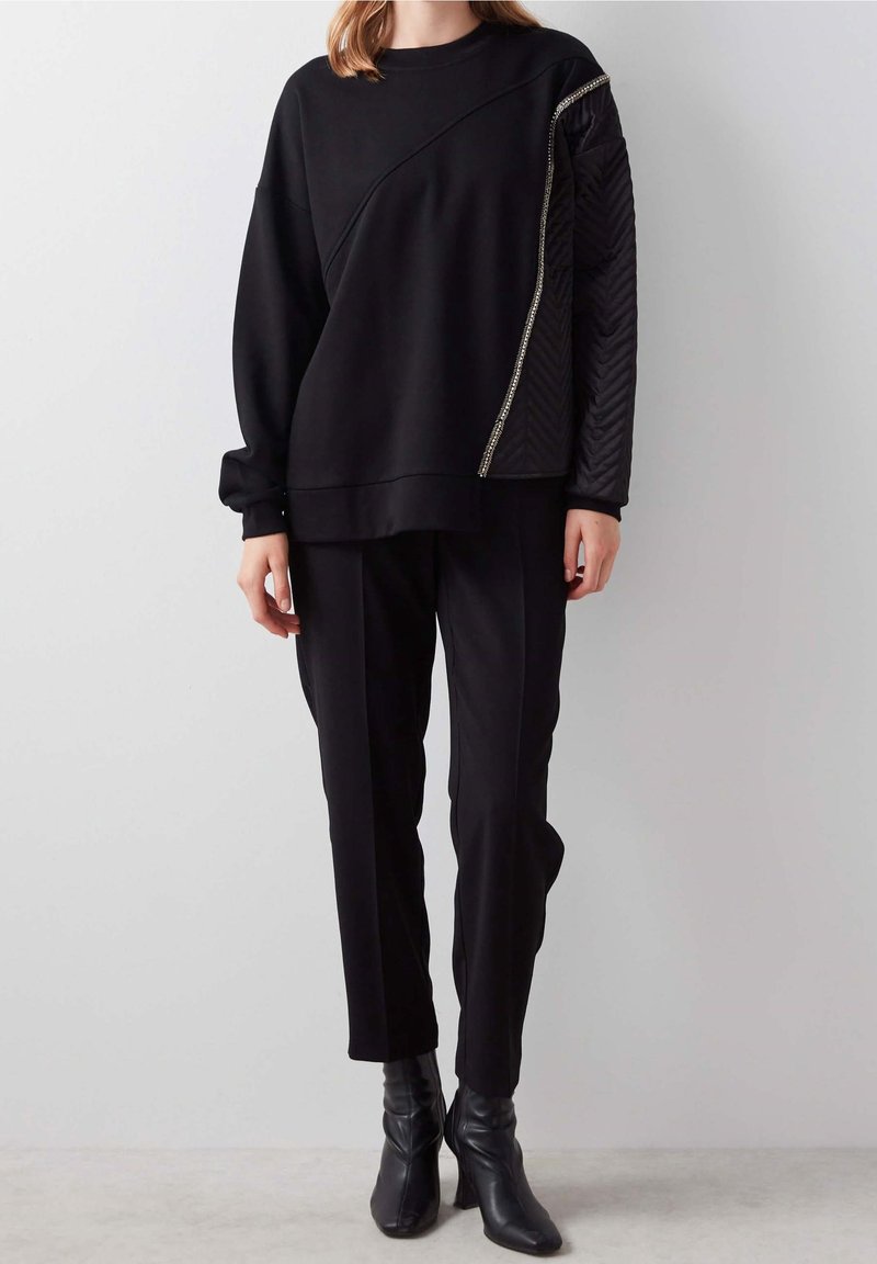 Personne portant un pantalon noir et un haut noir à manches longues avec une couture diagonale et une manche droite matelassée soulignée par une fermeture éclair métallique.