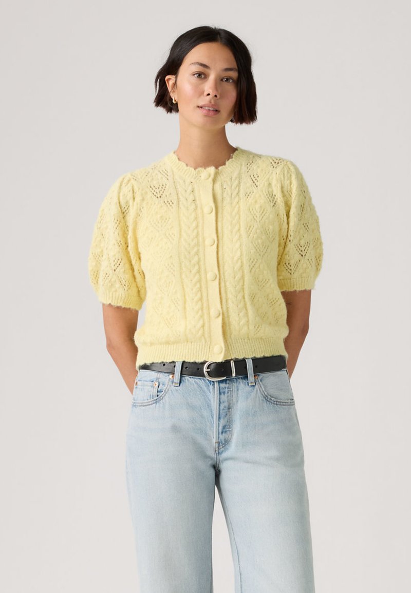 Woman standing wearing a pale yellow knit cardigan with puffed short sleeves and light blue high-waisted jeans with a black belt.