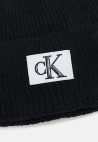 Gorra de lana negra con un dobladillo que presenta un parche blanco con las iniciales "cK" en una fuente negra brillante. Material suave y texturizado.