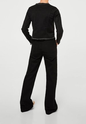 Personne portant une veste noire courte à manches longues et un pantalon large assorti, debout avec les mains dans les poches arrière devant un fond uni.