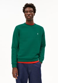 Felpa verde con scollatura rotonda, polsini a costine e un piccolo logo bianco a nastro sul petto, indossata sopra una camicia rossa. Pantaloni in denim blu.