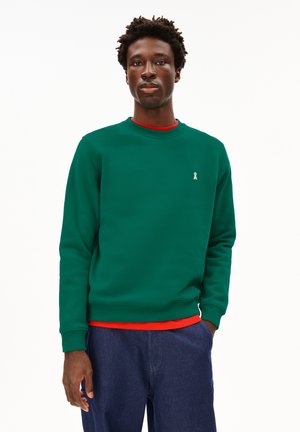 Groene sweatshirt met een ronde halslijn, geribbelde manchetten en een klein wit lintlogo op de borst, over een rode shirt. Blauwe spijkerbroek.
