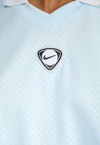 Gaiši zils polo krekls ar rūtainu rakstu, kuram uz krūšu zonas ir izšūts melns un balts Nike logo. Mīksta auduma tekstūra.