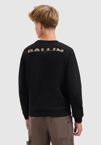 Tienerjongen met kort lichtbruin haar, gekleed in een zwart sweatshirt met de tekst "BALLIN" op de bovenrug en bruine cargobroeken.