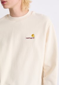 Krämfärgad sweatshirt i mjukt tyg med ribbat halsband och en liten broderad Carhartt-logga i brunt och gult på bröstet.