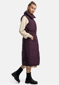 Gilet lungo senza maniche in viola scuro, con colletto alto, dettaglio della cintura e tasche laterali. Indossato con un maglione beige e stivali neri.
