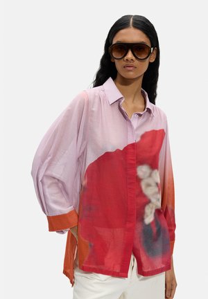 Kobieta w dużych brązowych okularach przeciwsłonecznych, luźnej koszuli w wielokolorowe odcienie różu, czerwieni i pomarańczy oraz białych spodniach, stojąca na jednolitym tle.