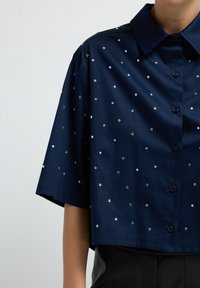 Camicetta blu navy a maniche corte con un design accorciato, caratterizzata da abbellimenti metallici a forma di quadrato e una chiusura frontale con bottoni. Tessuto dalla texture liscia.