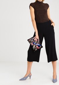 Kobieta w czarnych, szerokich spodniaczkach, brązowej bluzce z krótkim rękawem, szarych szpilkach, trzymająca czarną torebkę z kwiatowym haftem.