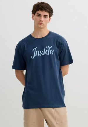 Hombre joven vestido con camiseta azul marino con texto "Inside" y pantalones beige, de pie frente a un fondo liso de color gris claro.