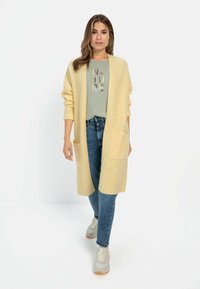 Geel gebreide cardigan met een open voorkant en zakken, gedragen over een lichtgroene T-shirt met de tekst "NATURE", gecombineerd met blauwe jeans en sneakers.