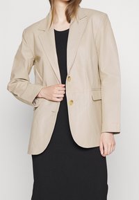 Bežová kožená blazer se zářezovými klopami, dvěma zlatými knoflíky a bočními kapsami; hladká textura; nošená přes černé žebrované šaty.