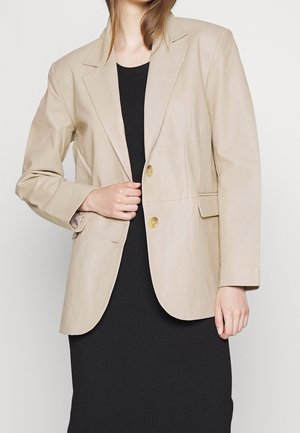 Blazer in pelle beige con rever a punta, due bottoni dorati e tasche laterali; texture liscia; indossato su un vestito nero a coste.
