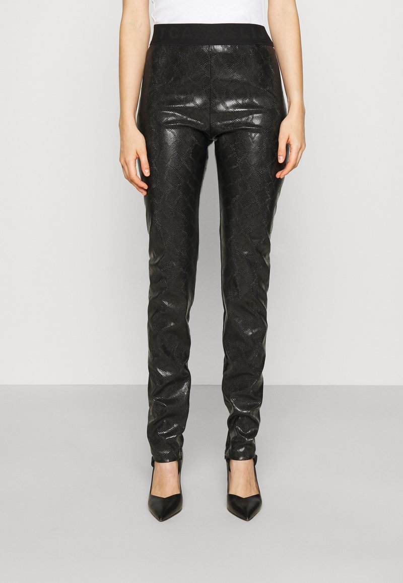 Pantalon en similicuir noir avec un motif texturé effet peau de serpent, coupe ajustée, et une ceinture élastique noire unie. Porté avec des talons pointus noirs.