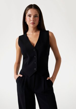Gilet sans manches noir avec un décolleté en V profond, fermeture à cinq boutons et une petite poche. Le tissu a une texture lisse ; conçu avec une coupe décontractée.