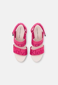 Růžové sandály s širokými popruhy, na nichž je logo "Calvin Klein". Podešev je hladké, světlé barvy. Součástí je také nastavitelný pásek na patě.