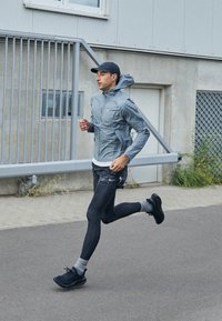 Muž v šedé bundě, černé čepici a běžeckých botách běží po dlažbě vedle průmyslové budovy s kovovou bránou a betonovou zdí.