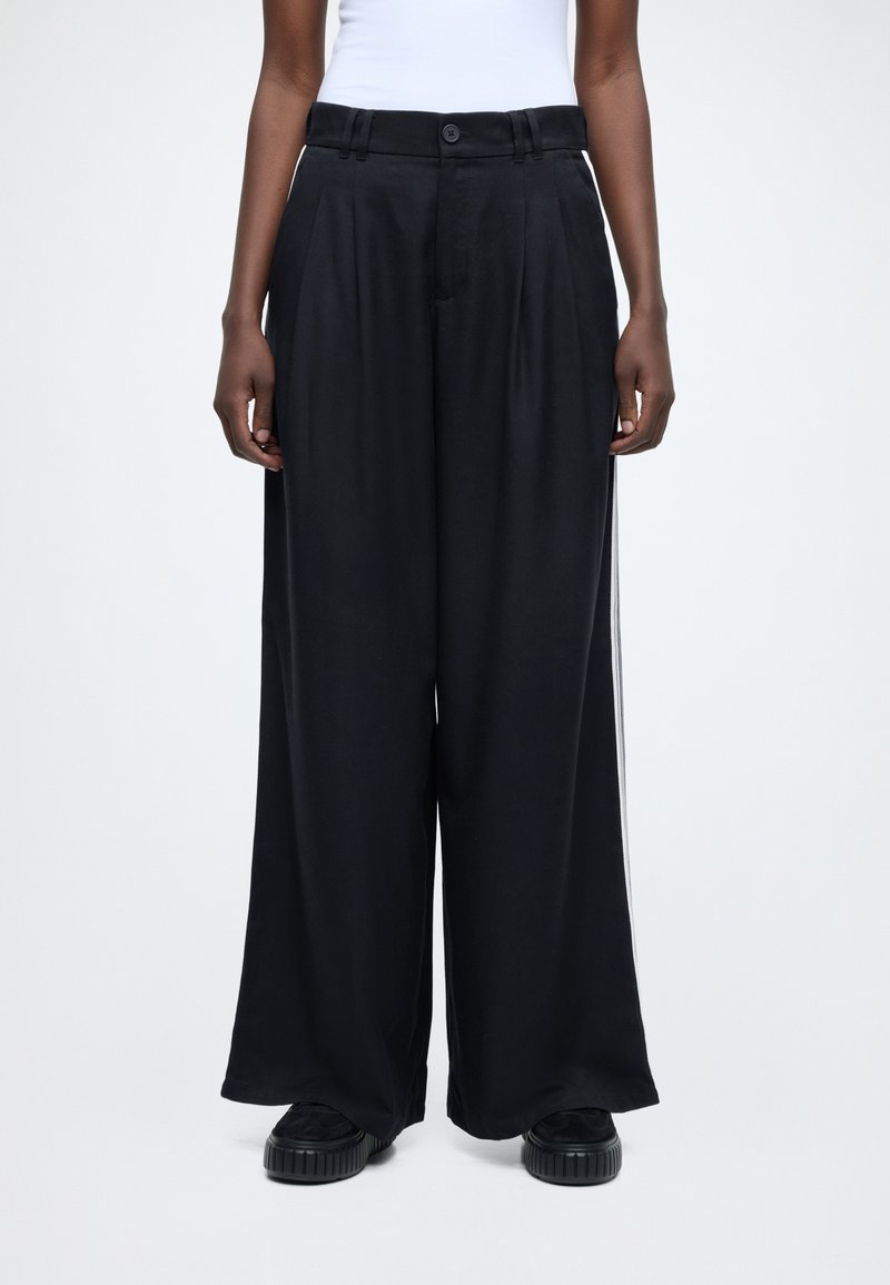 Črne široke hlače z gladko teksturo, visokim pasom, sprednjimi gubami in zapenjanjem na gumbe, kombinirane z teksturiranimi črnimi platformnimi čevlji.